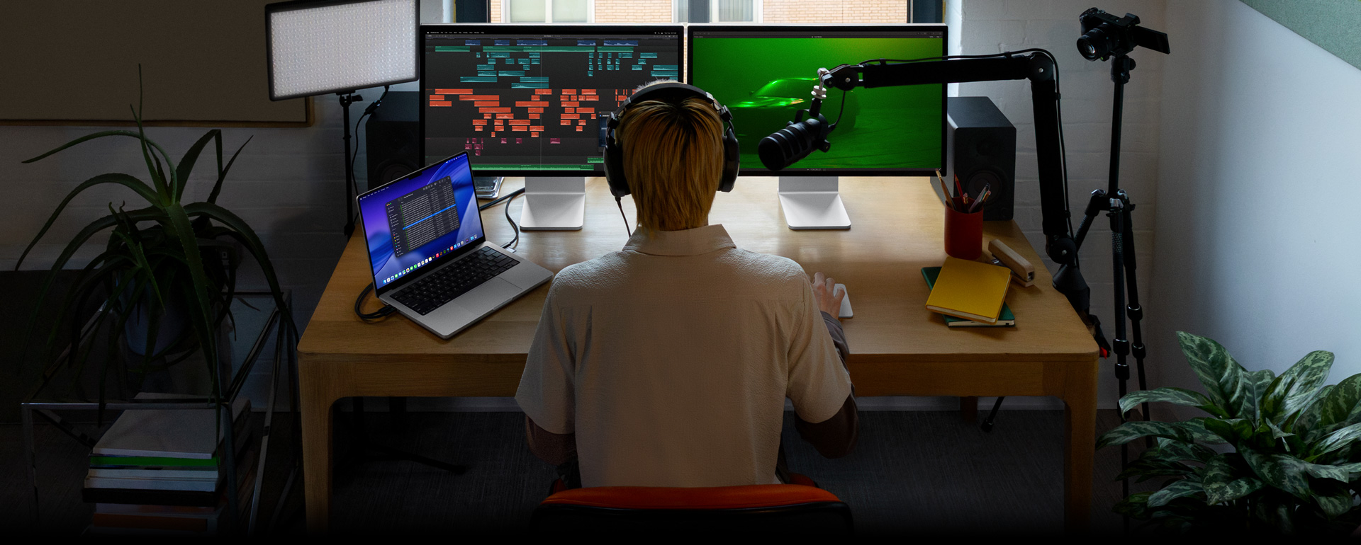 Person sitting at desk with mic and headphones in front of three devices, Mac Studio Display showing detailed audio editing tools, sound clips in different blocks of colour, a second Mac Studio Display screen showing car with moody green lighting, MacBook Pro 16 screen long list of shots in a Final Cut Pro project