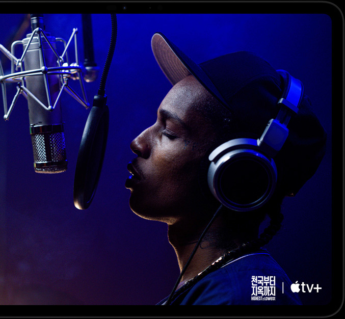 가로 방향으로 놓인 스페이스 블랙 색상 iPad Pro의 앞면. 둥근 형태의 모서리, 블랙 디스플레이 베젤이 보이고 화면에는 Apple TV 오리지널 영화 '천국부터 지옥까지' - Highest 2 Lowest에 등장하는 Yung Felon 역의 에이셉 라키가 마이크 앞에서 랩을 하고 있는 모습이 재생되고 있습니다.