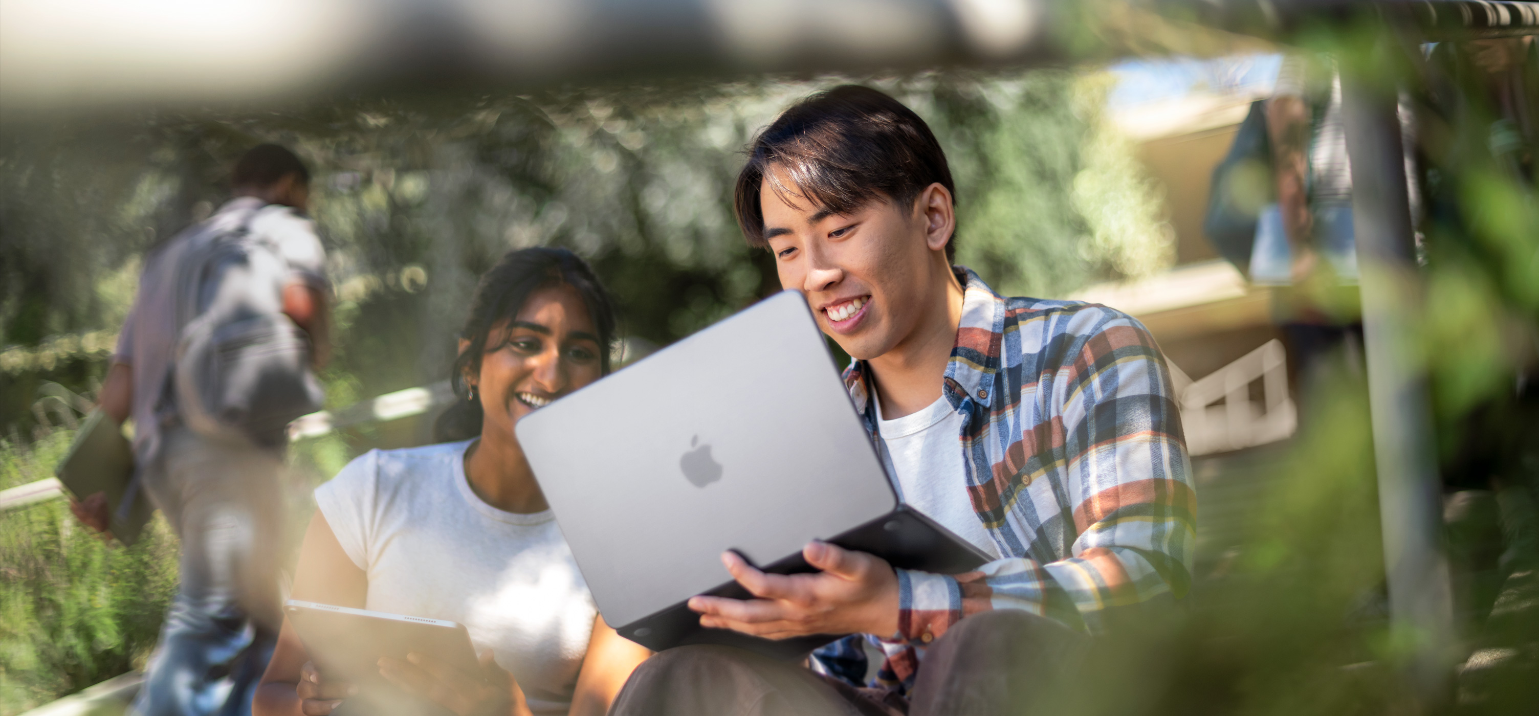 Two smiling students use MacBook outdoors, one holds it on their lap as they collaborate on an assignment