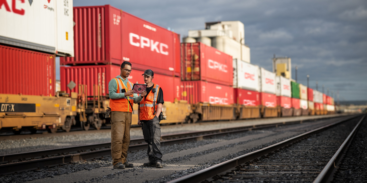 Två järnvägsarbetare står på en bangård bredvid ett stillastående tåg fullastat med containrar och tittar på en iPad