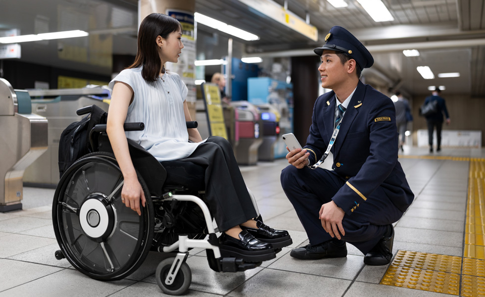 A professionally dressed woman in a wheelchair and a station attendant have an exchange on the platform.
