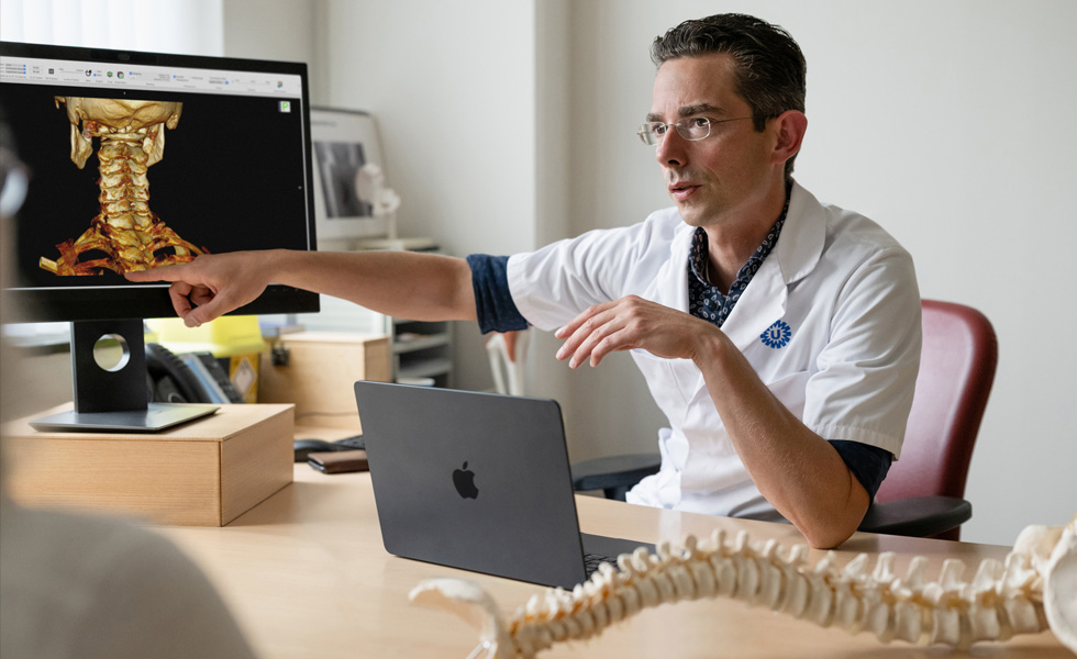 A medical professional points to a 3D skeletal rendering of the back of a neck on a Studio&nbsp;Display, while a MacBook sits on the desk