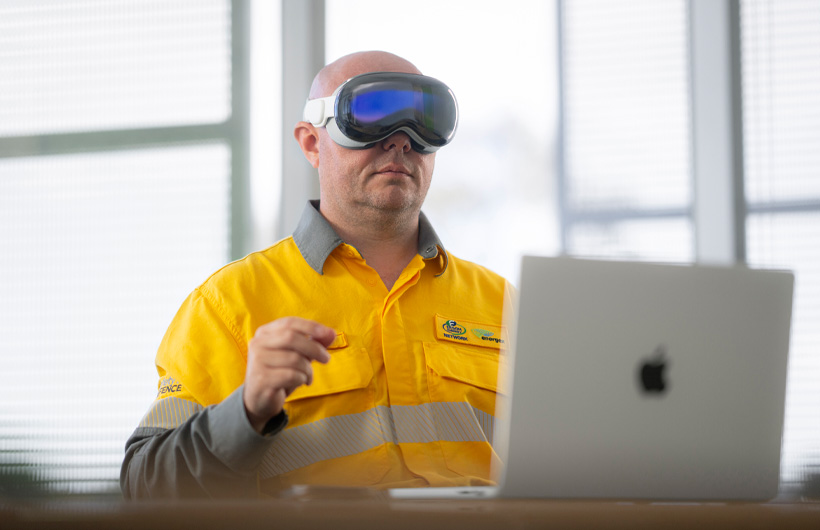An employee wears Apple Vision Pro and makes a pinching gesture with their hands, while a MacBook Pro in Silver sits open on the desk