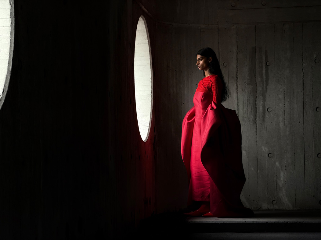 A woman wearing a long red dress stands in an empty room being photographed. The Photos app toggles through the focal lengths to demonstrate the capabilities of the pro camera system, showcasing: 100mm