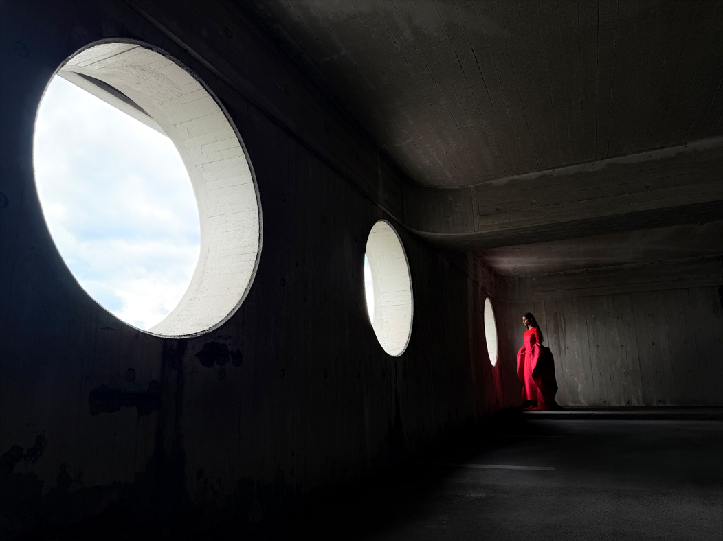 A woman wearing a long red dress stands in an empty room being photographed. The Photos app toggles through the focal lengths to demonstrate the capabilities of the pro camera system, showcasing: 24mm