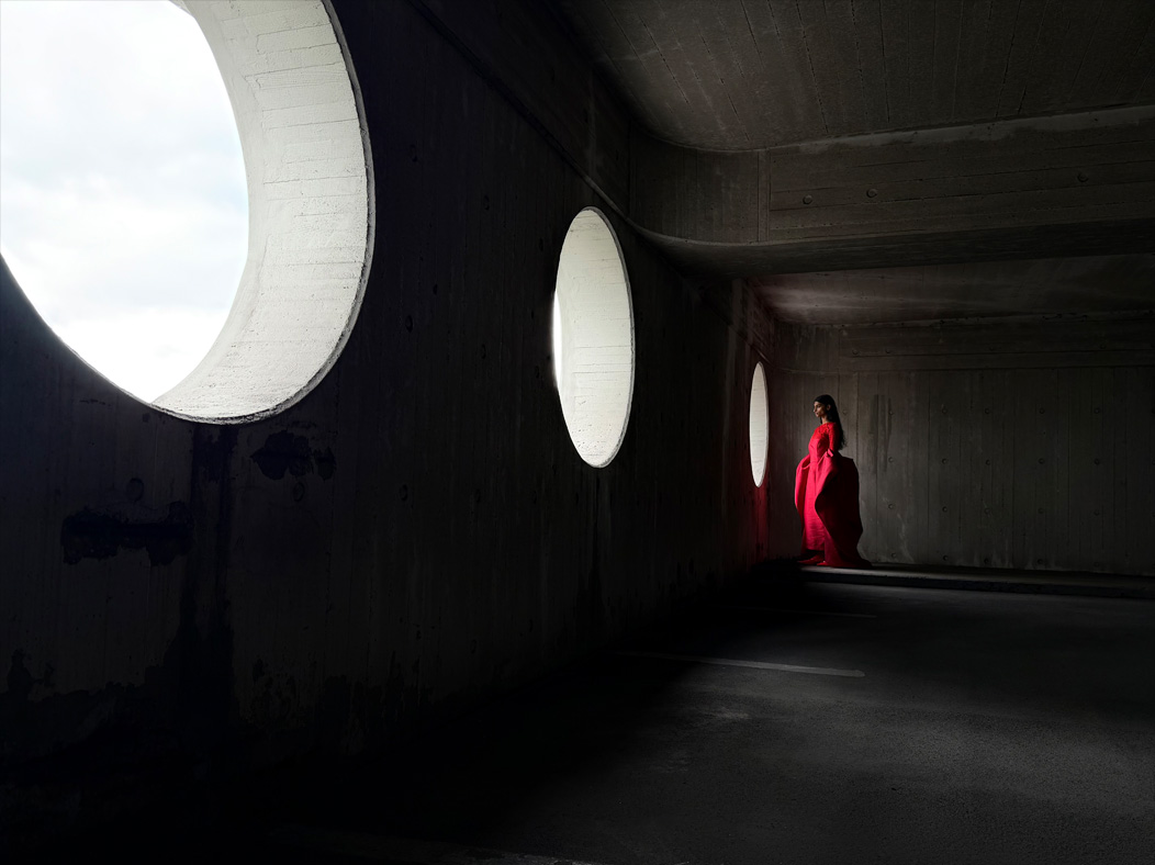 A woman wearing a long red dress stands in an empty room being photographed. The Photos app toggles through the focal lengths to demonstrate the capabilities of the pro camera system, showcasing: 28mm