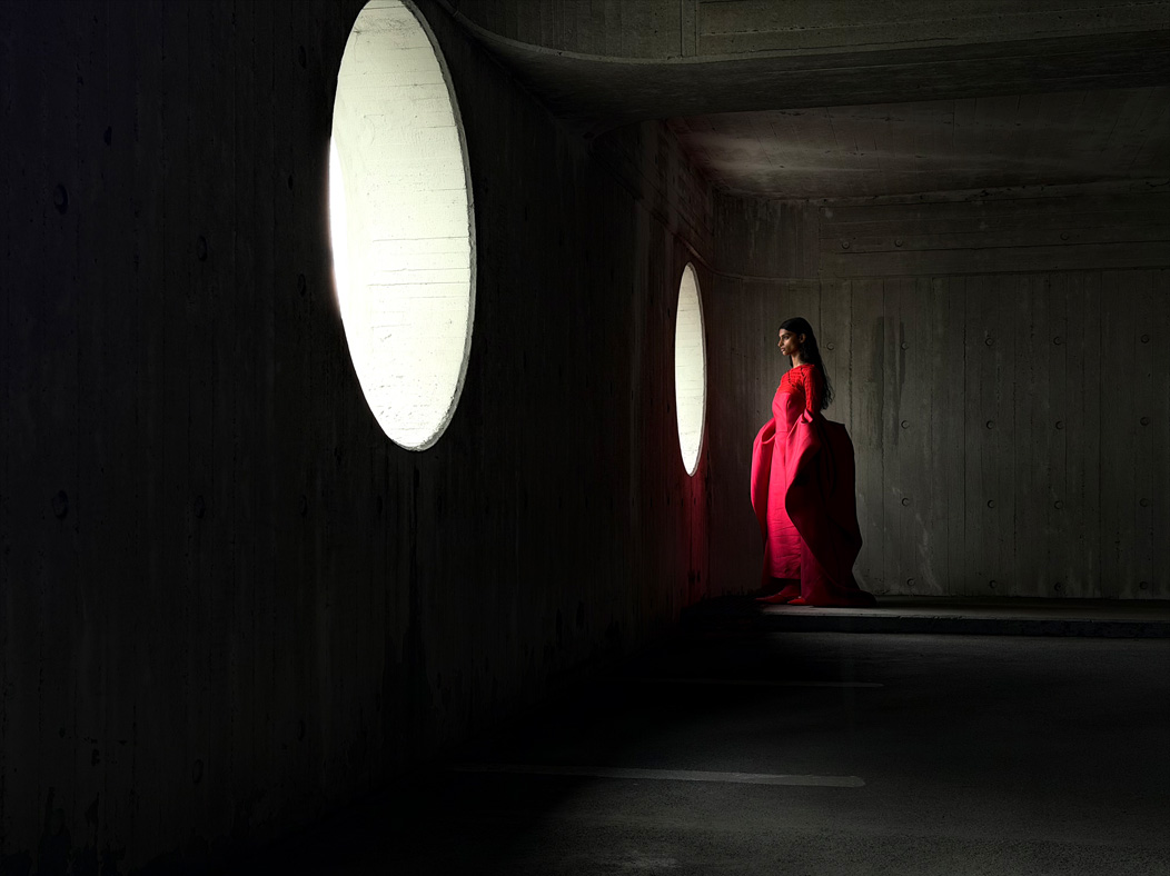 A woman wearing a long red dress stands in an empty room being photographed. The Photos app toggles through the focal lengths to demonstrate the capabilities of the pro camera system, showcasing: 48mm