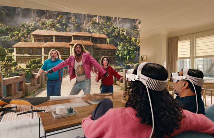 Apple Vision Pro를 착용한 두 사람이 소파에 나란히 앉아 방 전체에 펼쳐진 화면에 표시된 마인크래프트 무비를 보고 있는 모습